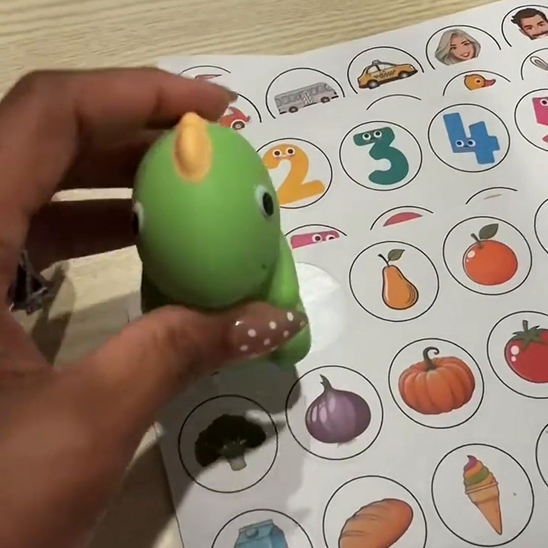 Real footage: a hand holds the green dino over a sticker sheet of animals, numbers and food, cards stacked behind.