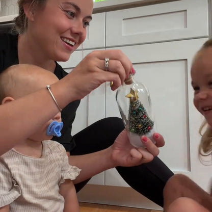 A mom with a baby and two toddlers on a rug, the bottle held in front of them