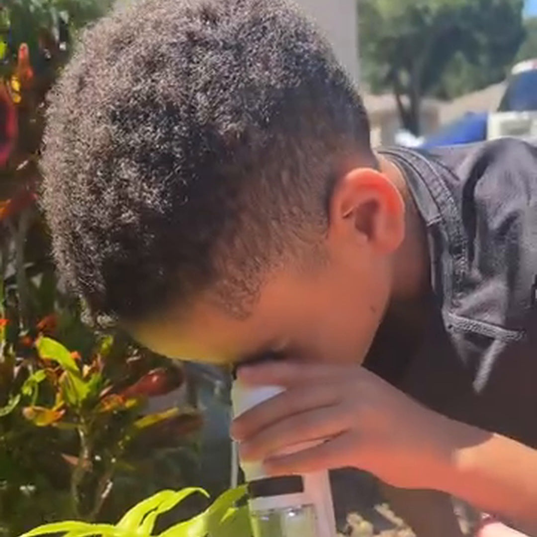 real frame: a boy crouched among green plants holding the microscope to his eye, no caption in the frame