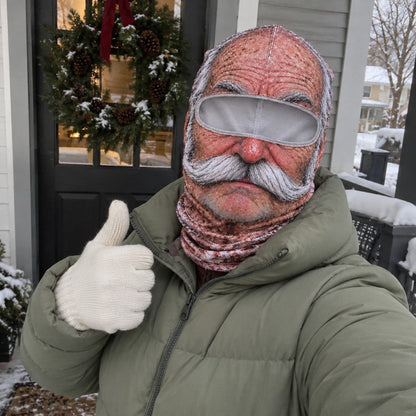 An adult in a sage puffer coat wearing the Grandpa mask on a snowy porch with a wreath.
