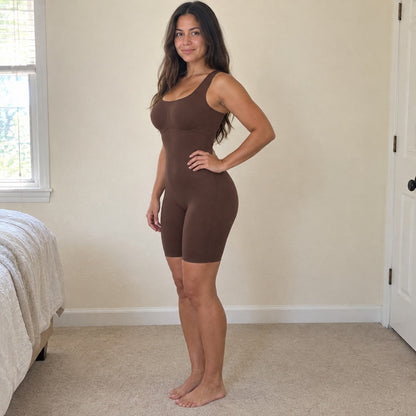 A woman stands side-on in a sunlit bedroom wearing the brown bodysuit, hand on hip, smooth tummy and lifted butt, cream walls set off the brown.