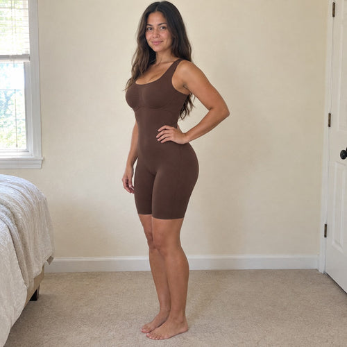 A woman stands side-on in a sunlit bedroom wearing the brown bodysuit, hand on hip, smooth tummy and lifted butt, cream walls set off the brown.