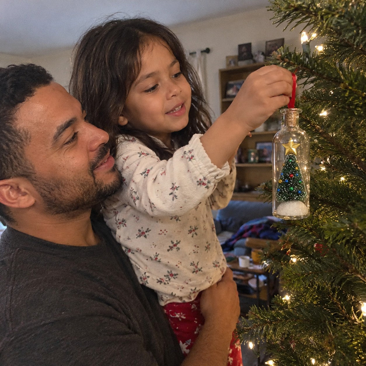 A father holding his daughter as she hangs one matching bottle by its red loop on a tree branch