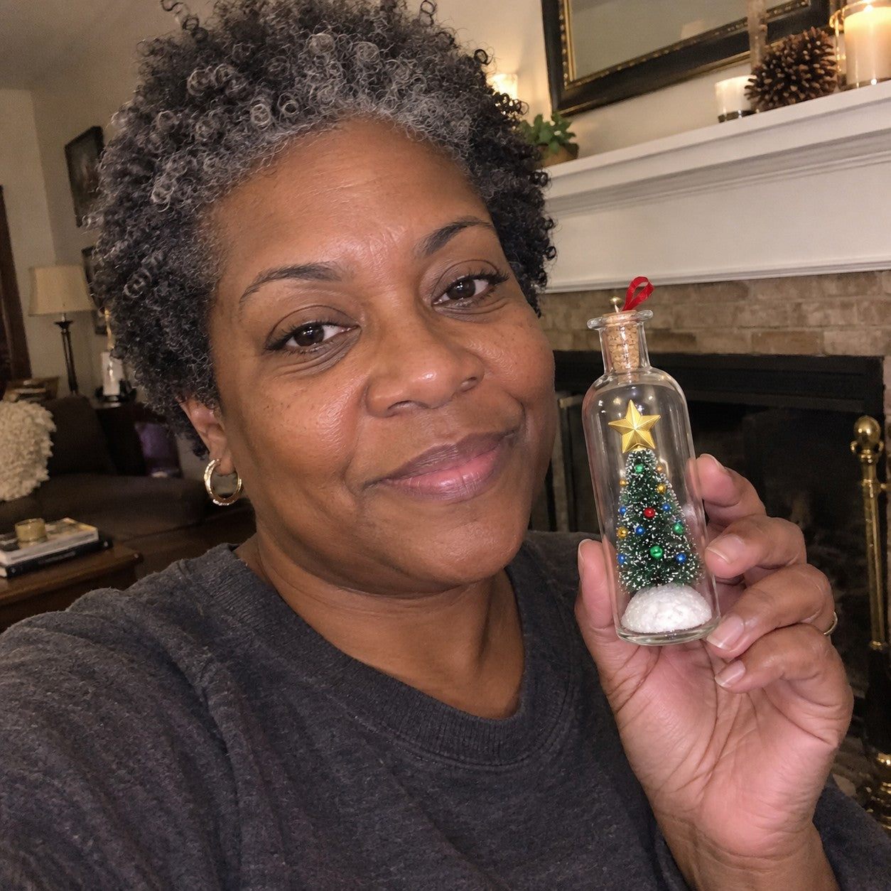 Denise C., a middle-aged woman with brown skin and short gray curls, holding one matching bottle beside her fireplace
