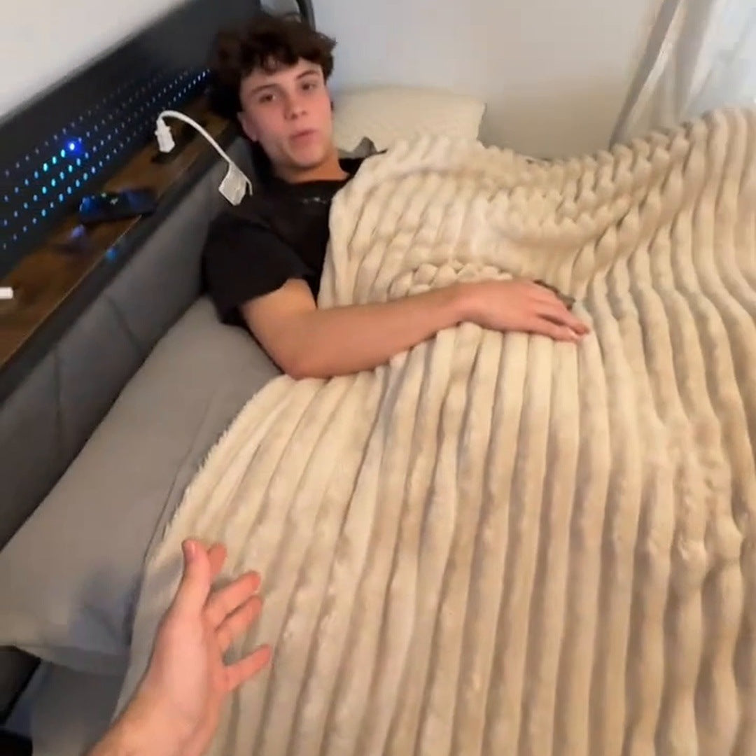 A young man in a black tee lying on a bed under the Cream ribbed blanket, a hand reaching in from the camera side