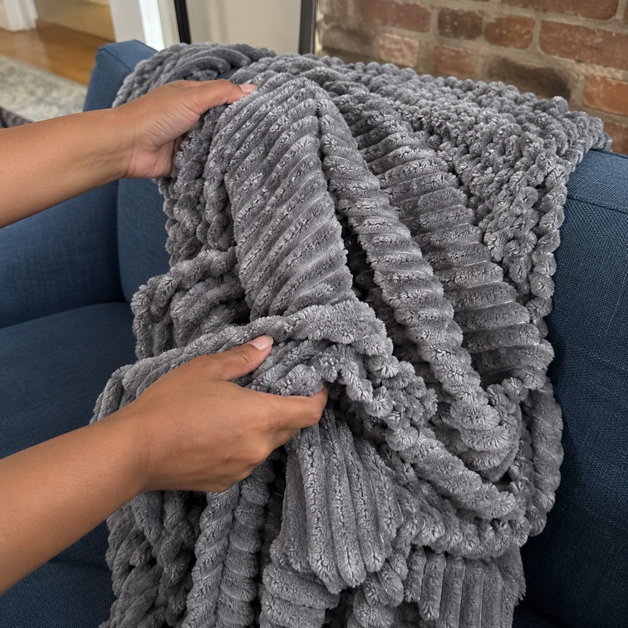 Maya hands gathering the Grey ribbed plush blanket on her blue couch