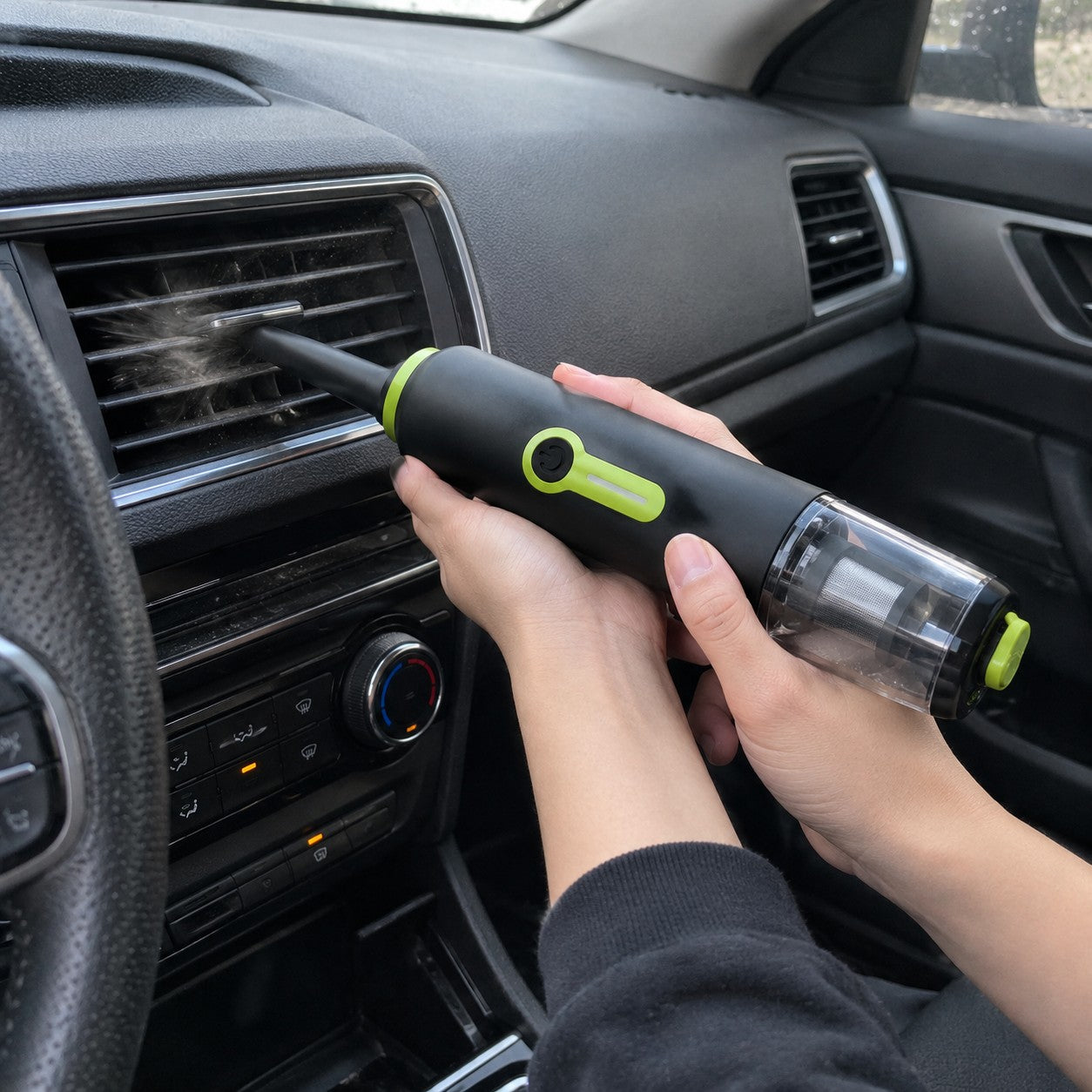 A hand points the rear blower nozzle at dashboard vent dust, with the clear cup at the opposite end