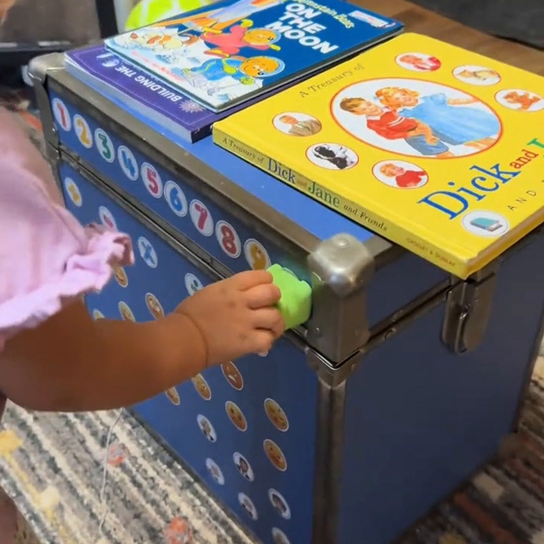 A little girl in a pink top taps the green dinosaur to a sticker on a blue trunk