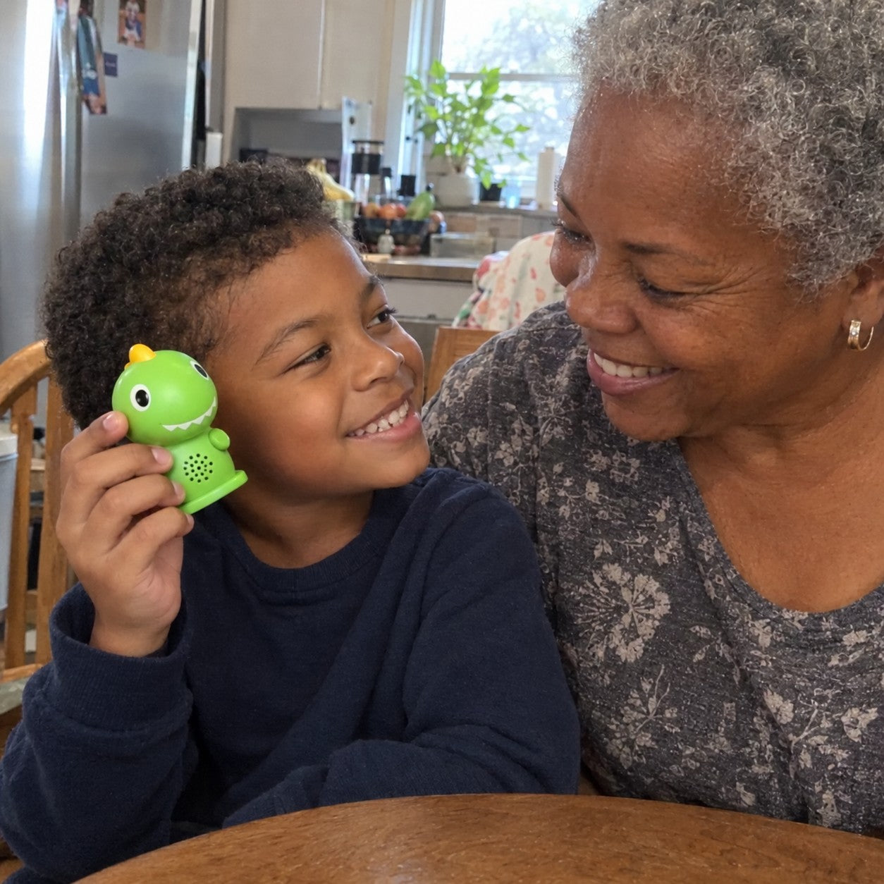A grandmother and her young grandson smile together as the boy holds the green dinosaur reader beside his face