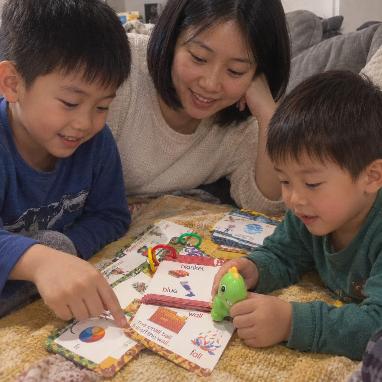 A mother watches her two boys take turns with the green dinosaur and illustrated word cards