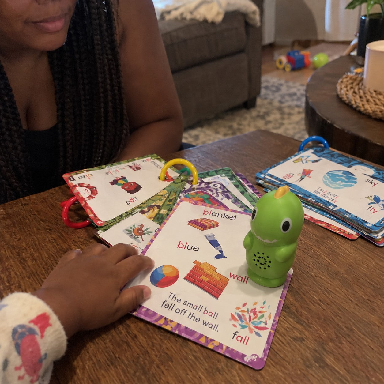 A mother with braids sits beside the green dinosaur and printed cards as a small hand reaches toward them