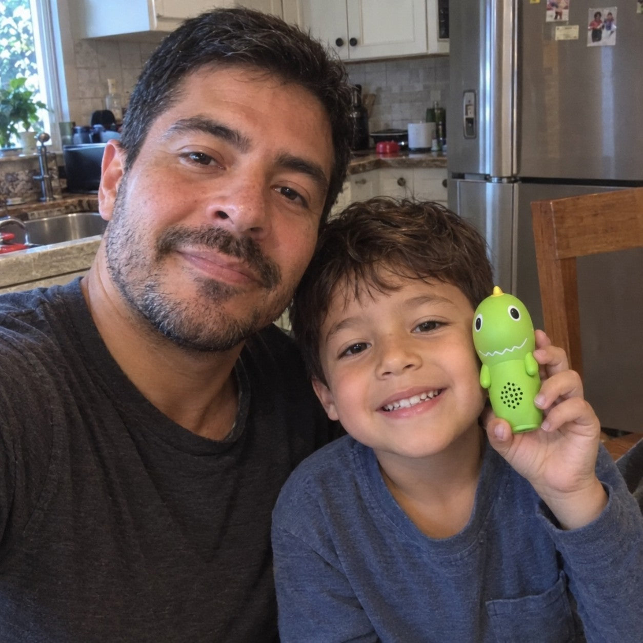 A dark-haired father beside his son, who holds the green dinosaur reader near his face