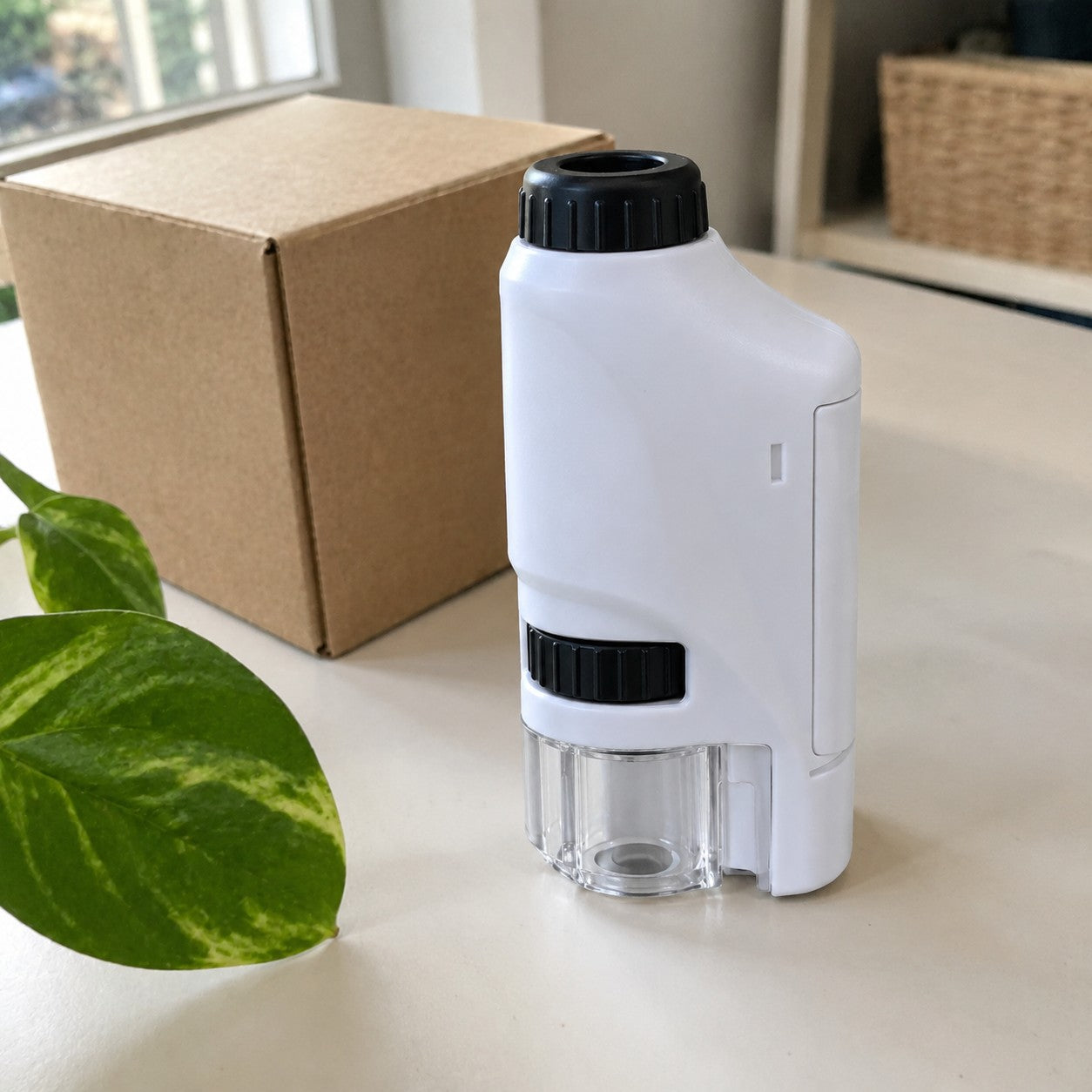 One white-and-black microscope stands on a pale table beside a closed plain shipping box and a green leaf