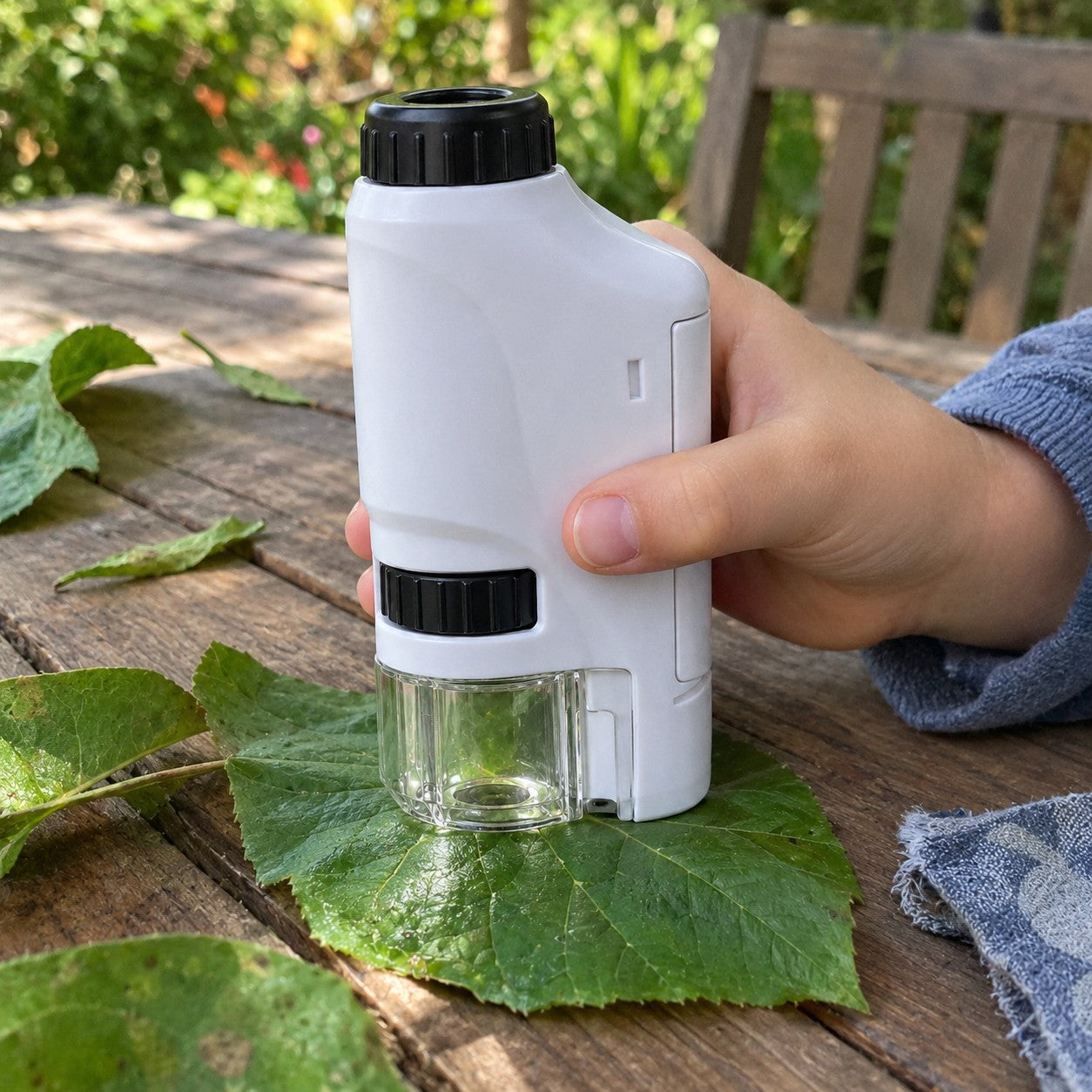 A small hand holds the white microscope onto a green leaf on an outdoor wooden table. Black controls and clear shroud match the reference