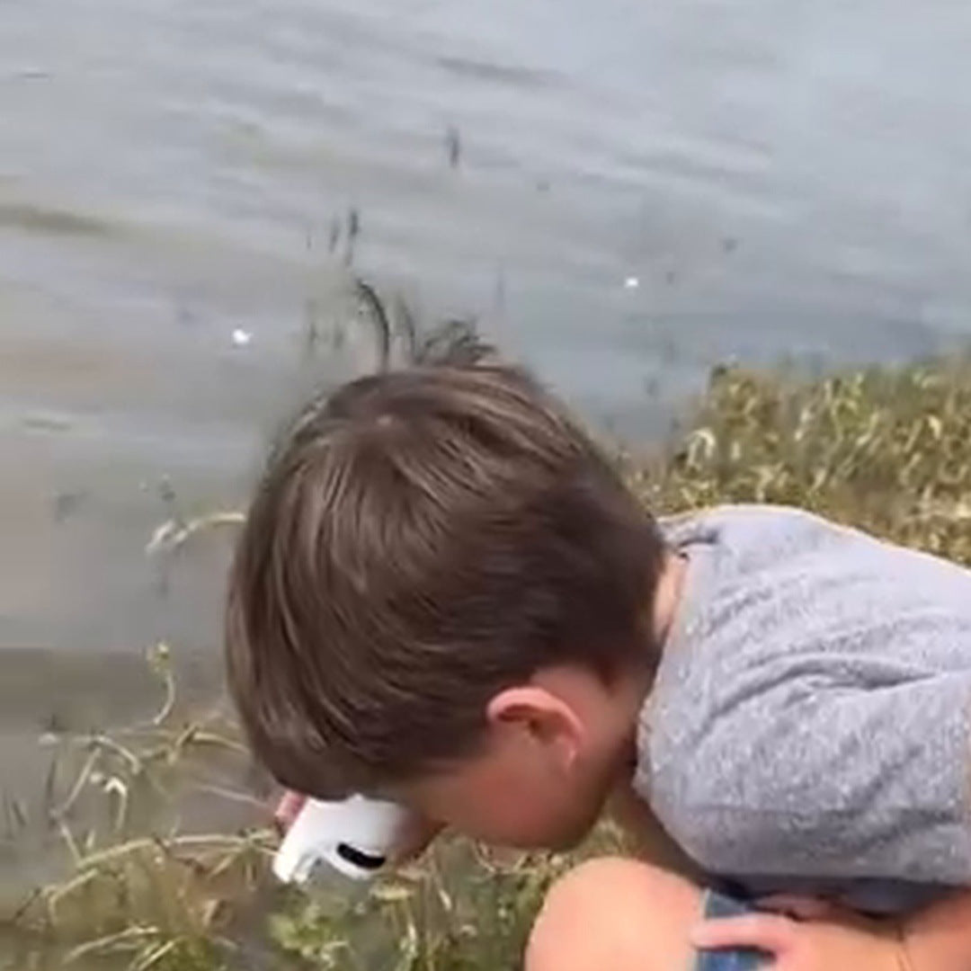 Real creative frame: a boy in a gray shirt bends over grass beside a lake with the microscope at his eye