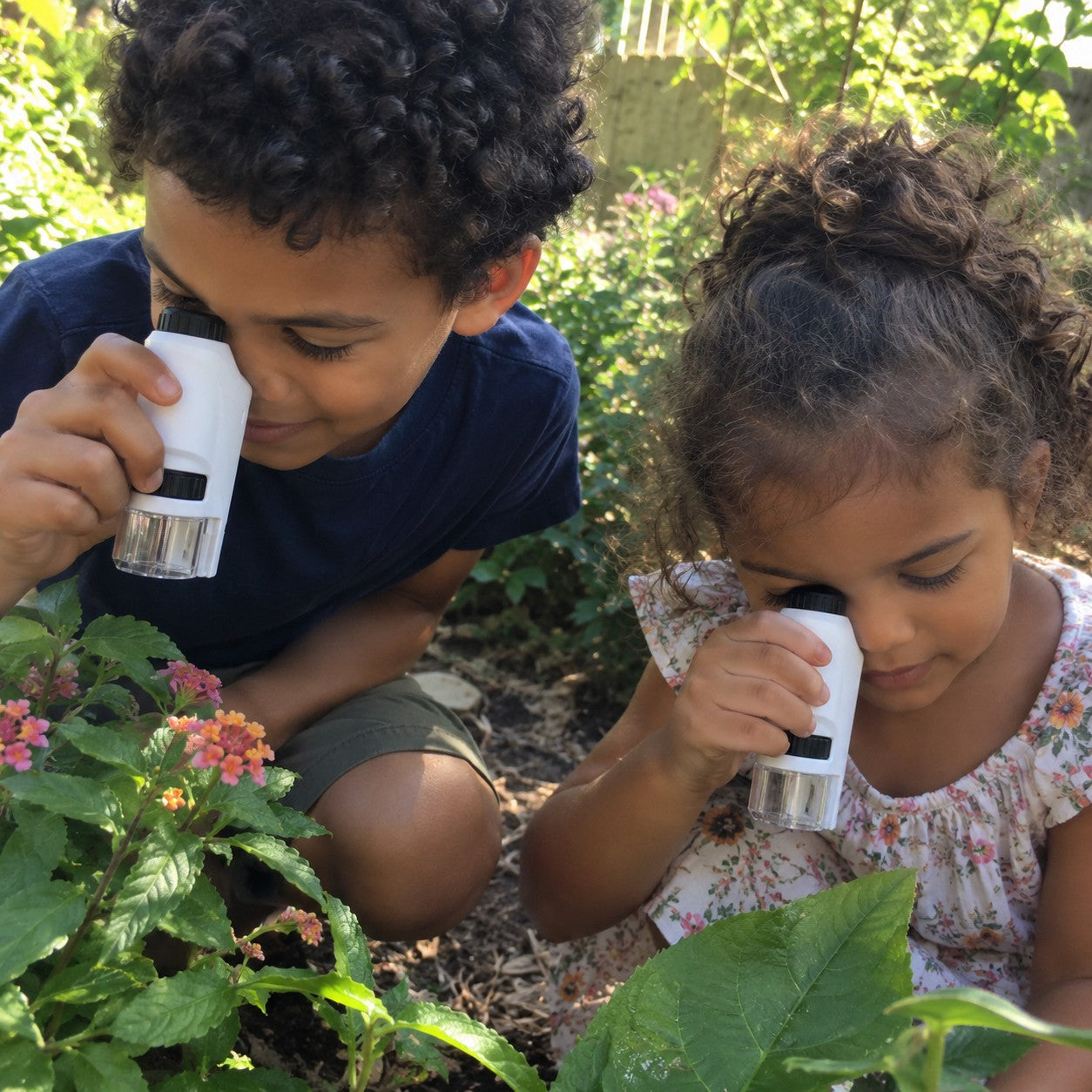 Two children each hold their own white-and-black microscope over garden flowers. This illustrates separate gifts and shared outdoor exploration