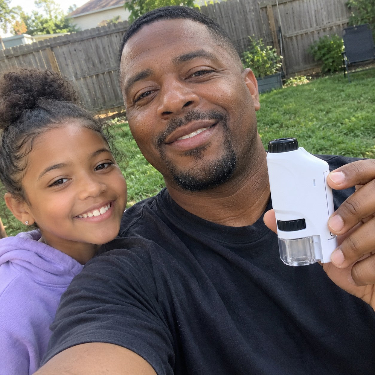 A father with short black hair and a goatee takes a backyard selfie with his daughter, holding the white microscope