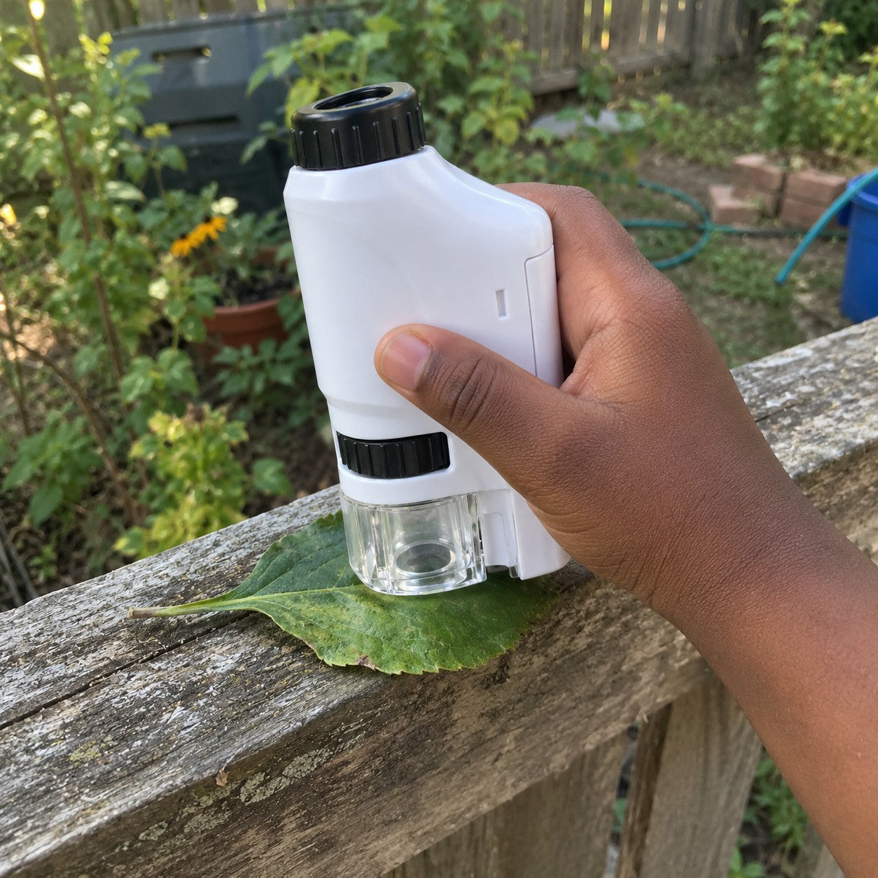 A dark-brown hand holds the white microscope over a leaf on a weathered garden fence ledge