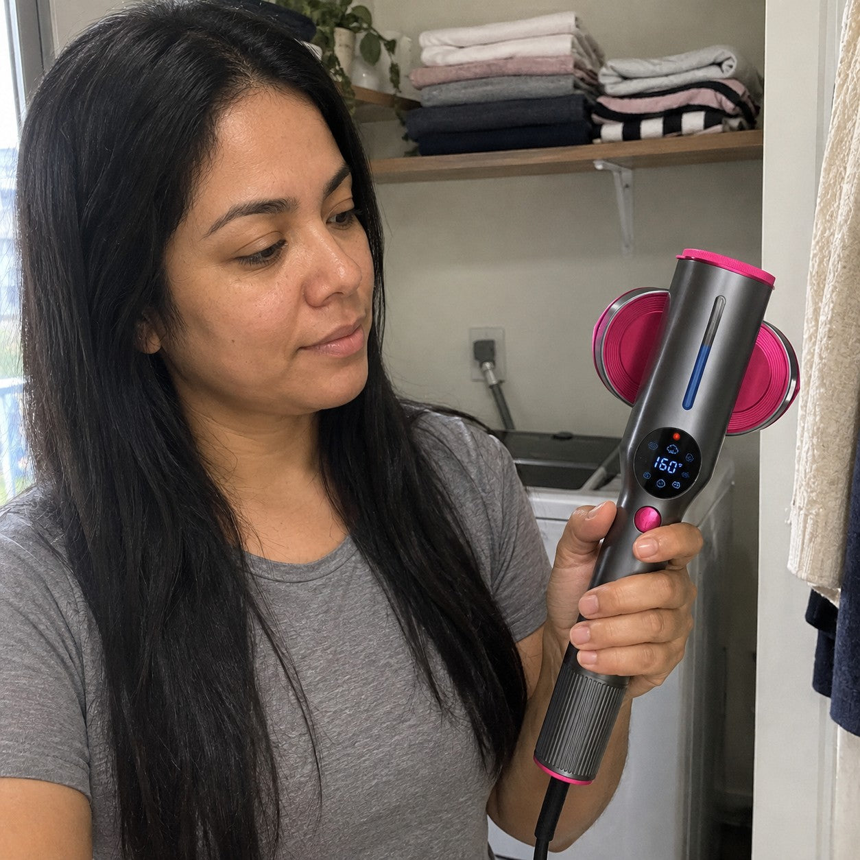 A woman in a laundry nook looks at the lit round display of the gray and magenta handheld held in her hand