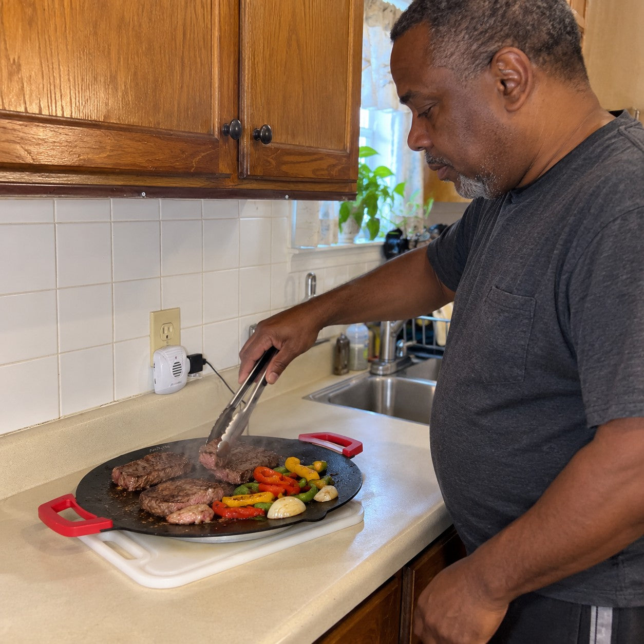 Marcus J.: deep brown skin and cropped black-gray hair, using tongs on steak and vegetables on the red-handled grill