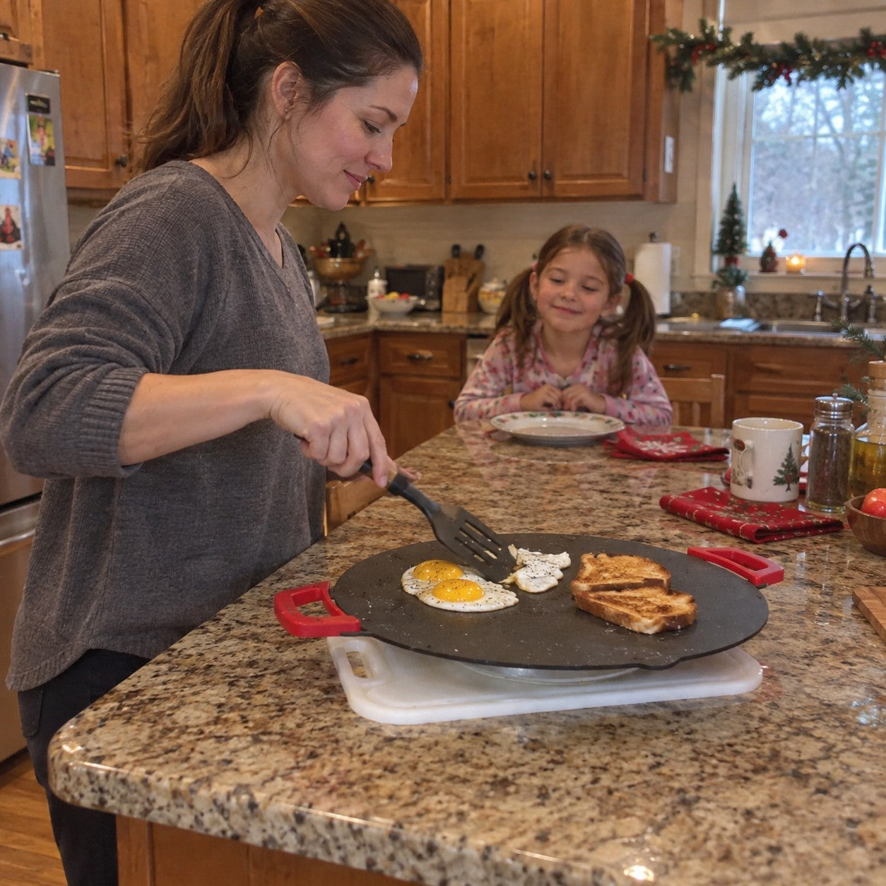 A mother cooks eggs and toast on the red-handled grill while her daughter stands back in a kitchen with a holiday garland