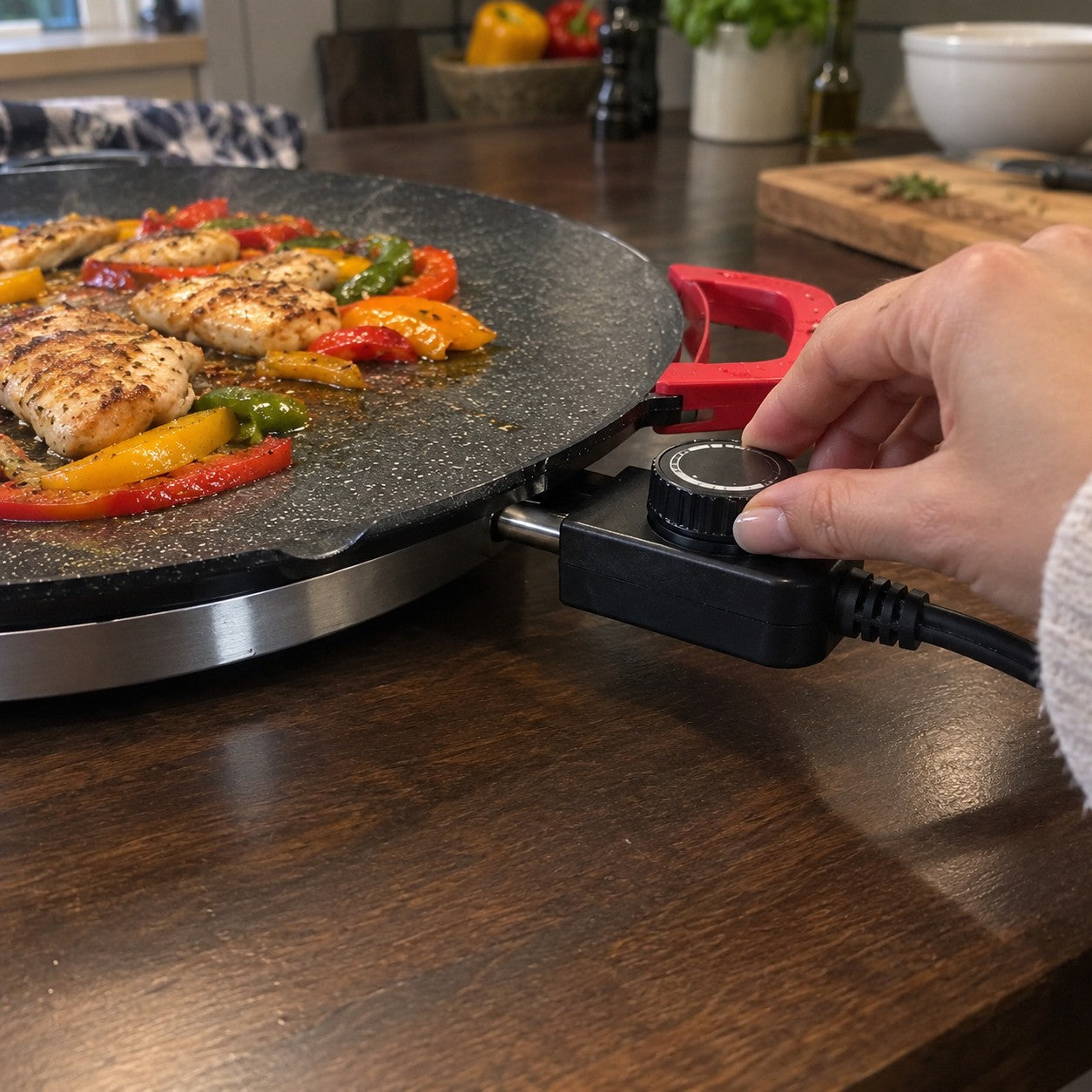 A hand turns the round black ridged temperature dial attached to the speckled grill, with chicken and peppers cooking
