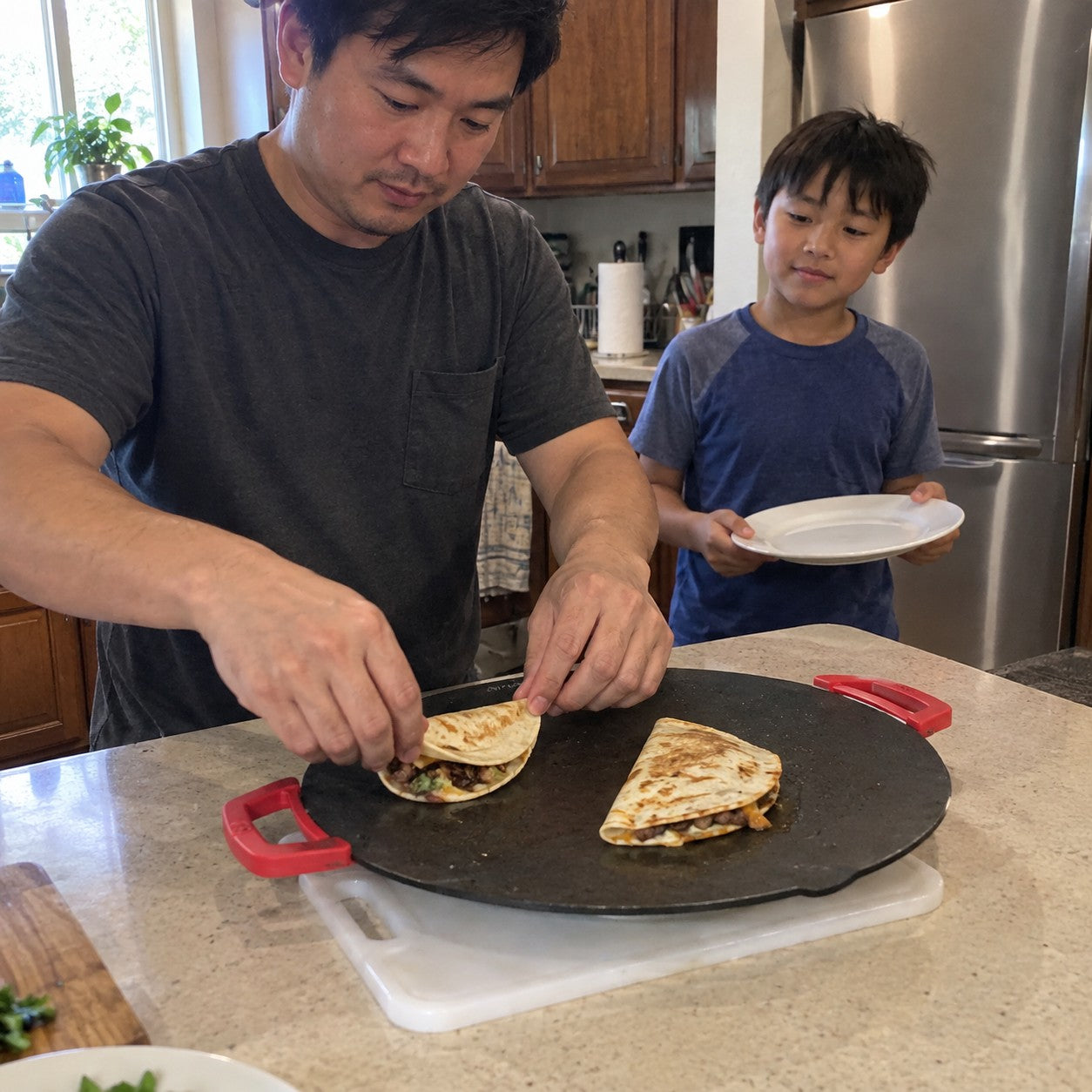 A father folds a tortilla on the grill while his son waits back with a plate in a home kitchen