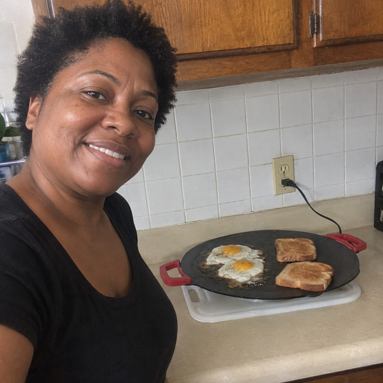 Denise M., short natural curls and warm brown skin, beside one red-handled grill cooking eggs and toast in a home kitchen