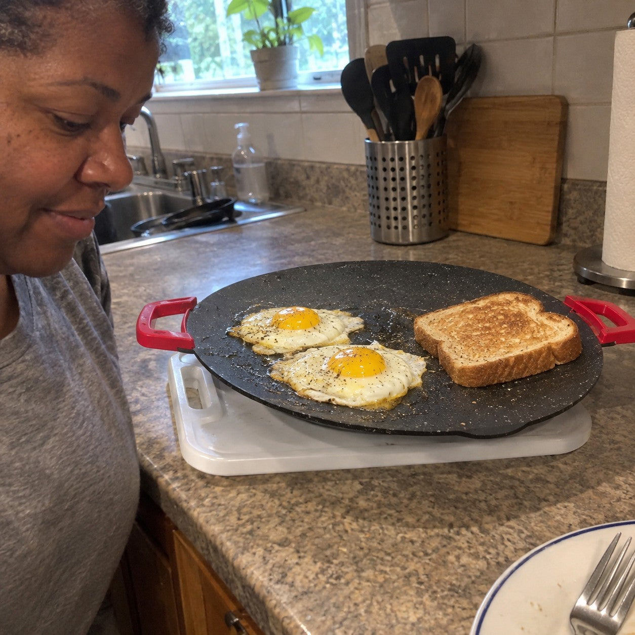 Denise M., short natural curls and warm brown skin, beside eggs and toast cooking on one red-handled grill
