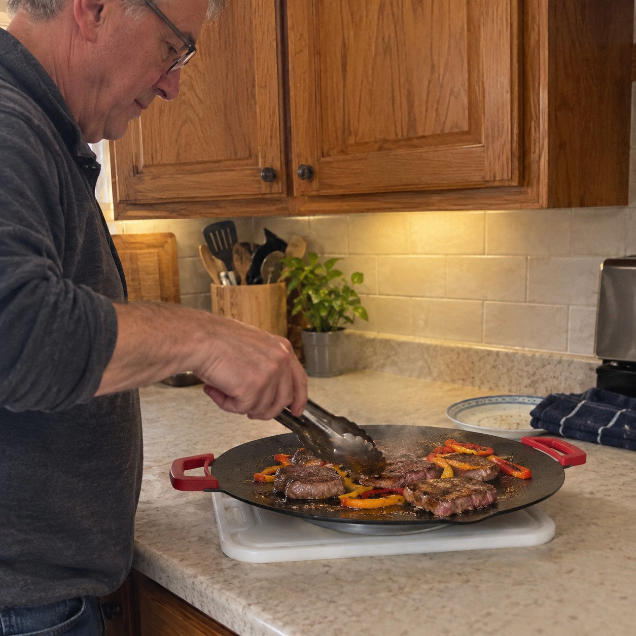 Tom H., gray hair and glasses, uses tongs to cook steak and peppers on one red-handled grill