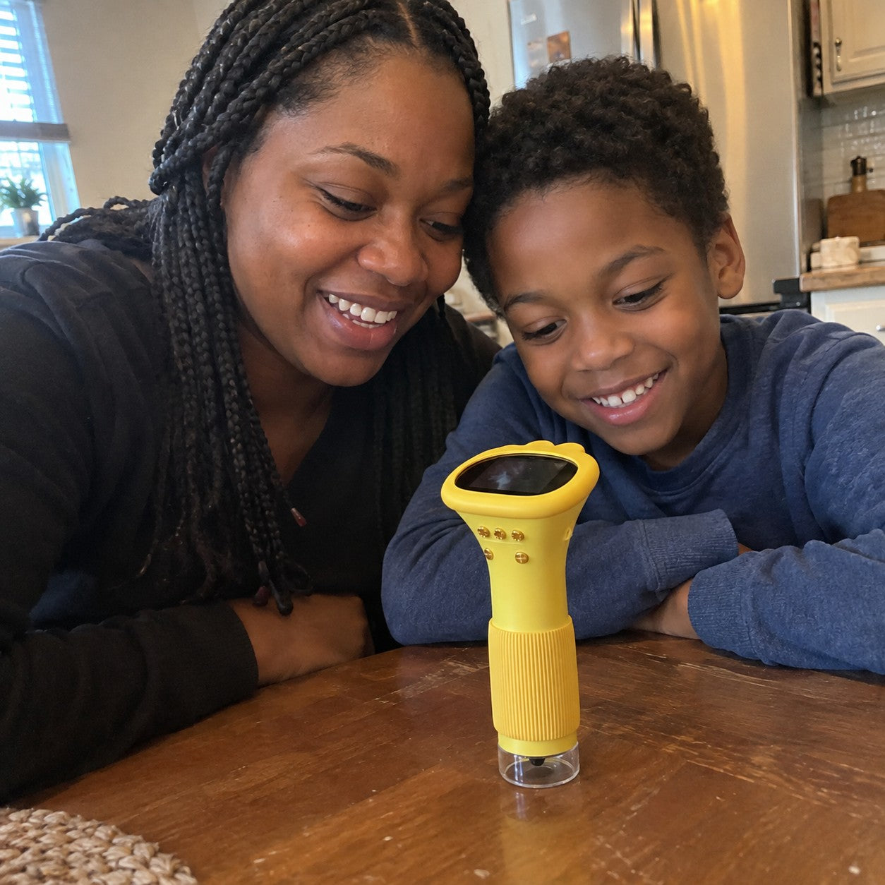 Mother and boy lean toward the shared yellow screen at a worn kitchen table