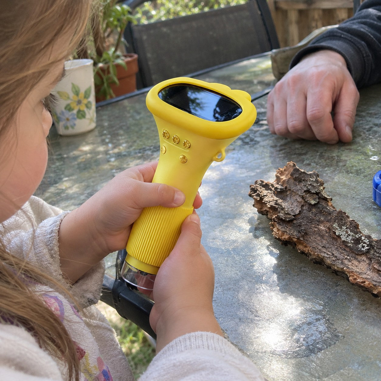 Child grips the yellow device at a patio table with bark and a nearby adult hand