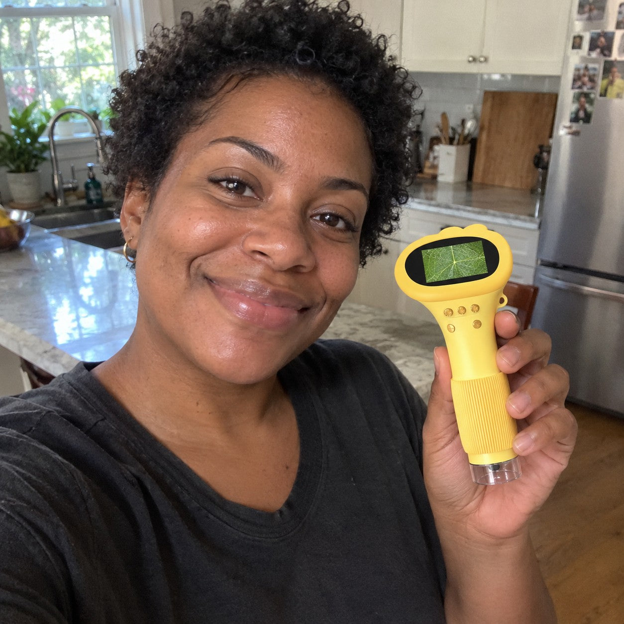 Dana W with natural curly black hair holds the yellow scope showing a leaf in a kitchen selfie